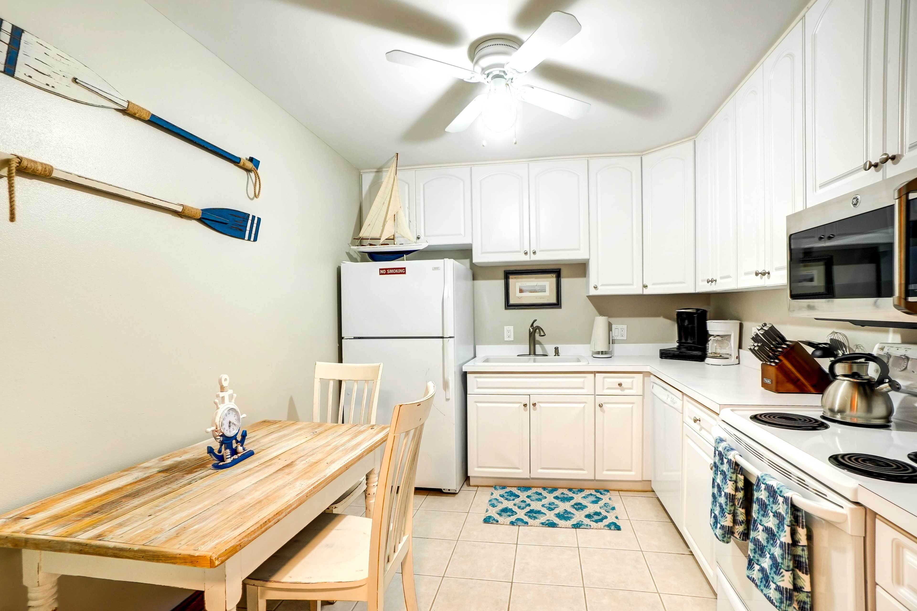 Une cuisine avec des meubles blancs, une table en bois, un ventilateur de plafond, un réfrigérateur, un four à micro-ondes et une bouilloire.