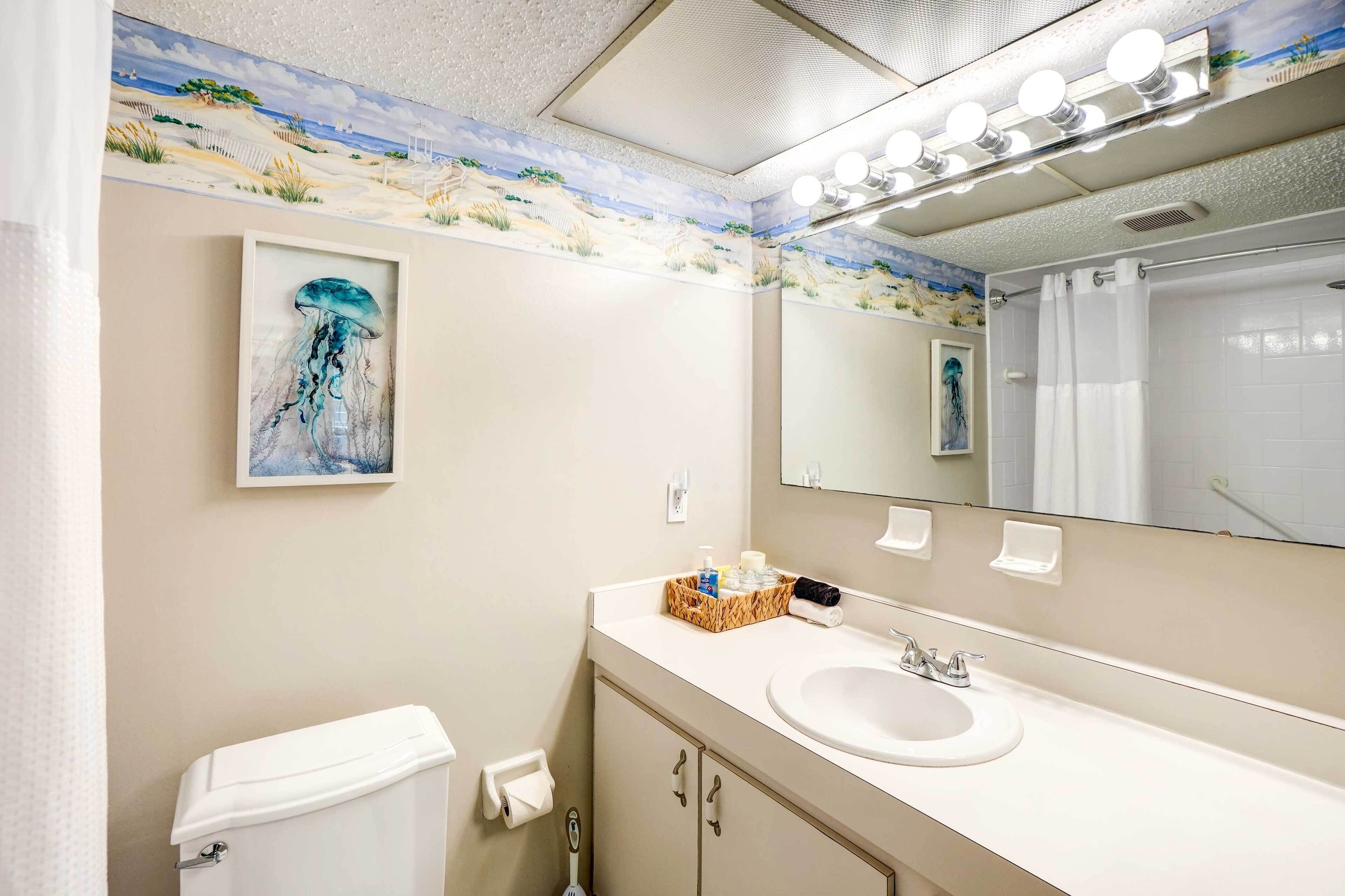 A bathroom with a sink, mirror, and toilet. There is a painting of a jellyfish on the wall and a beach scene above the mirror.