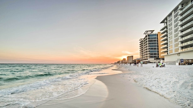A beach with a sandy shore and ocean waves, adjacent to a multi-story hotel with balconies and a clear view of the sunset. - Modern Resort Condo With Balcony - Walk to Beach! (Destin)