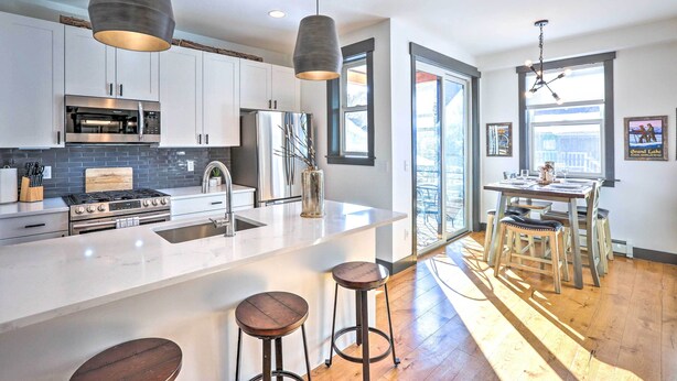 A modern kitchen with white cabinets, stainless steel appliances, a large island with stools, and a dining area with a view. - Modern Mountain Getaway, Walk to Grand Lake! (Grand Lake)