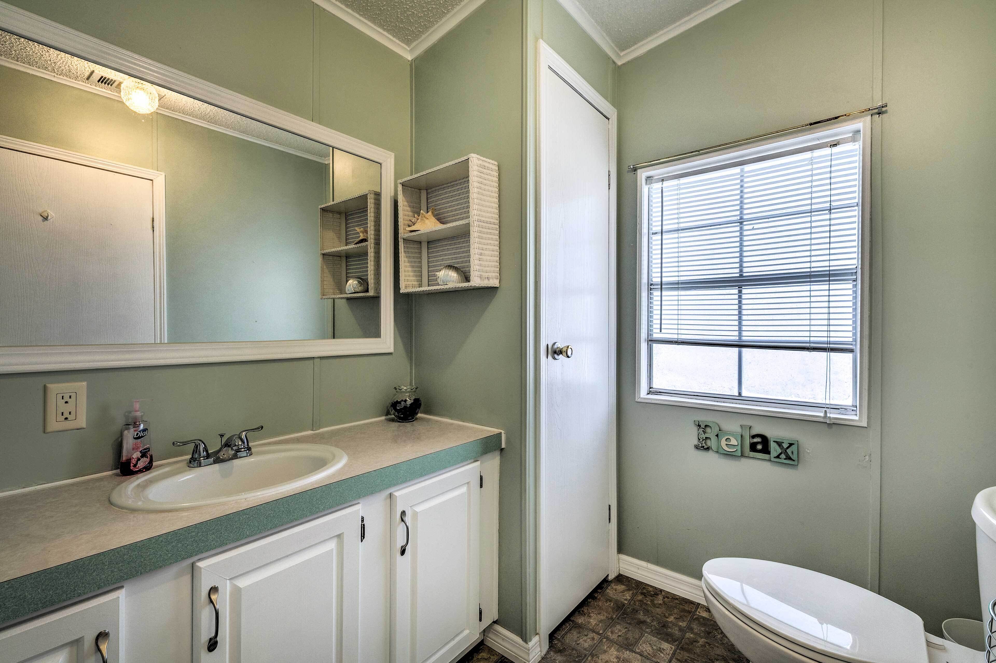 A bathroom with a sink, mirror, and toilet. There is a window with blinds and a shelf with toiletries.