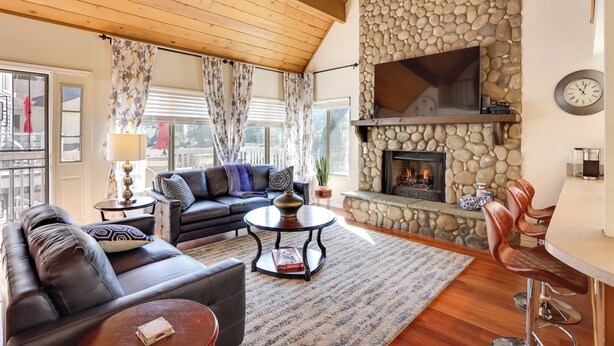 A cozy living room with a stone fireplace, leather furniture, and a large flat-screen TV. - Lake & Mountain-view Home: Walk to Downtown! (Crestline)