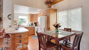 A modern kitchen with wooden cabinets, a central island with bar stools, and a dining area with a wooden table and chairs. - Lake & Mountain-view Home: Walk to Downtown! (Crestline)