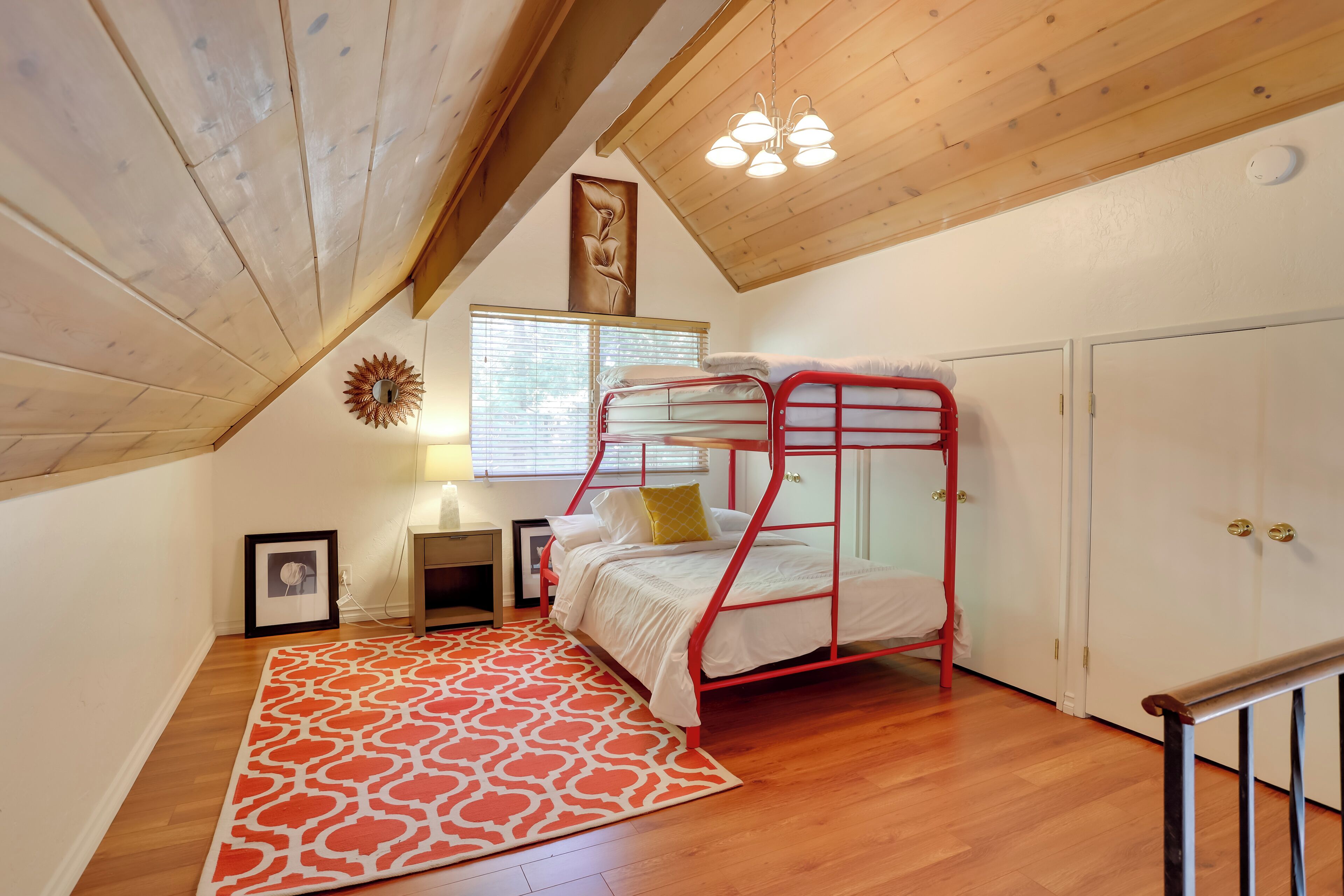 A dormitory room with a bunk bed, wooden flooring, a red and white patterned rug, a desk with a lamp, a framed picture, and a window with blinds.