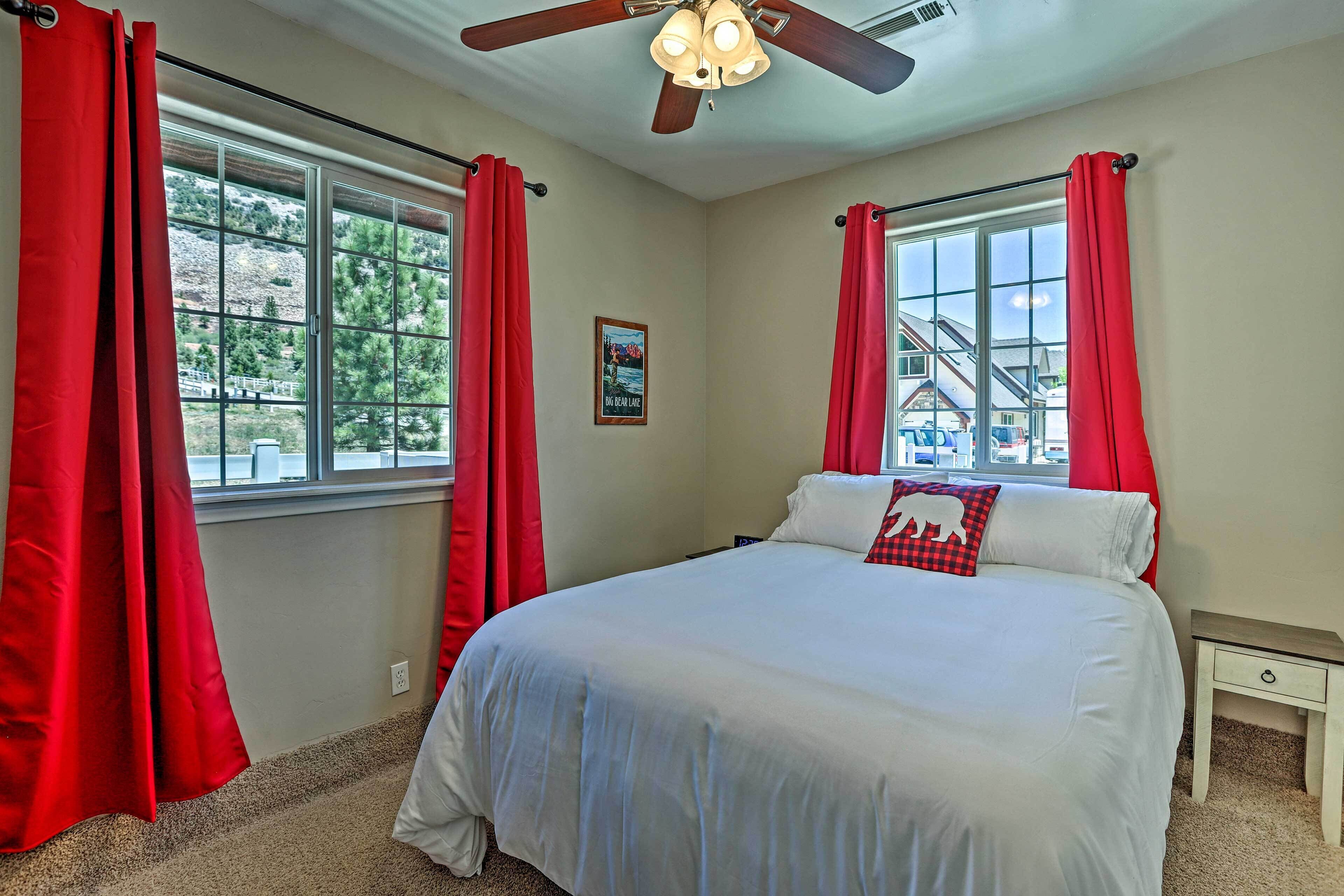 A bedroom with a large bed, red curtains, a ceiling fan, and a window with a view of a house and trees.