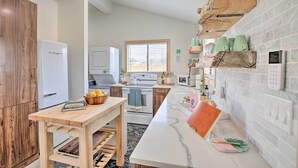 A modern kitchen with a wooden table, a white refrigerator, a stove, and a window with blinds. - Modern Joshua Tree Bungalow With Fire Pit & Bbq! (Joshua Tree)