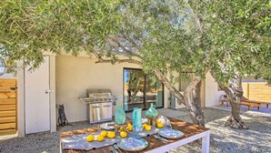 An outdoor dining area with a wooden table set for a meal, surrounded by olive trees, and a barbecue grill in the background. - Modern Joshua Tree Bungalow With Fire Pit & Bbq! (Joshua Tree)