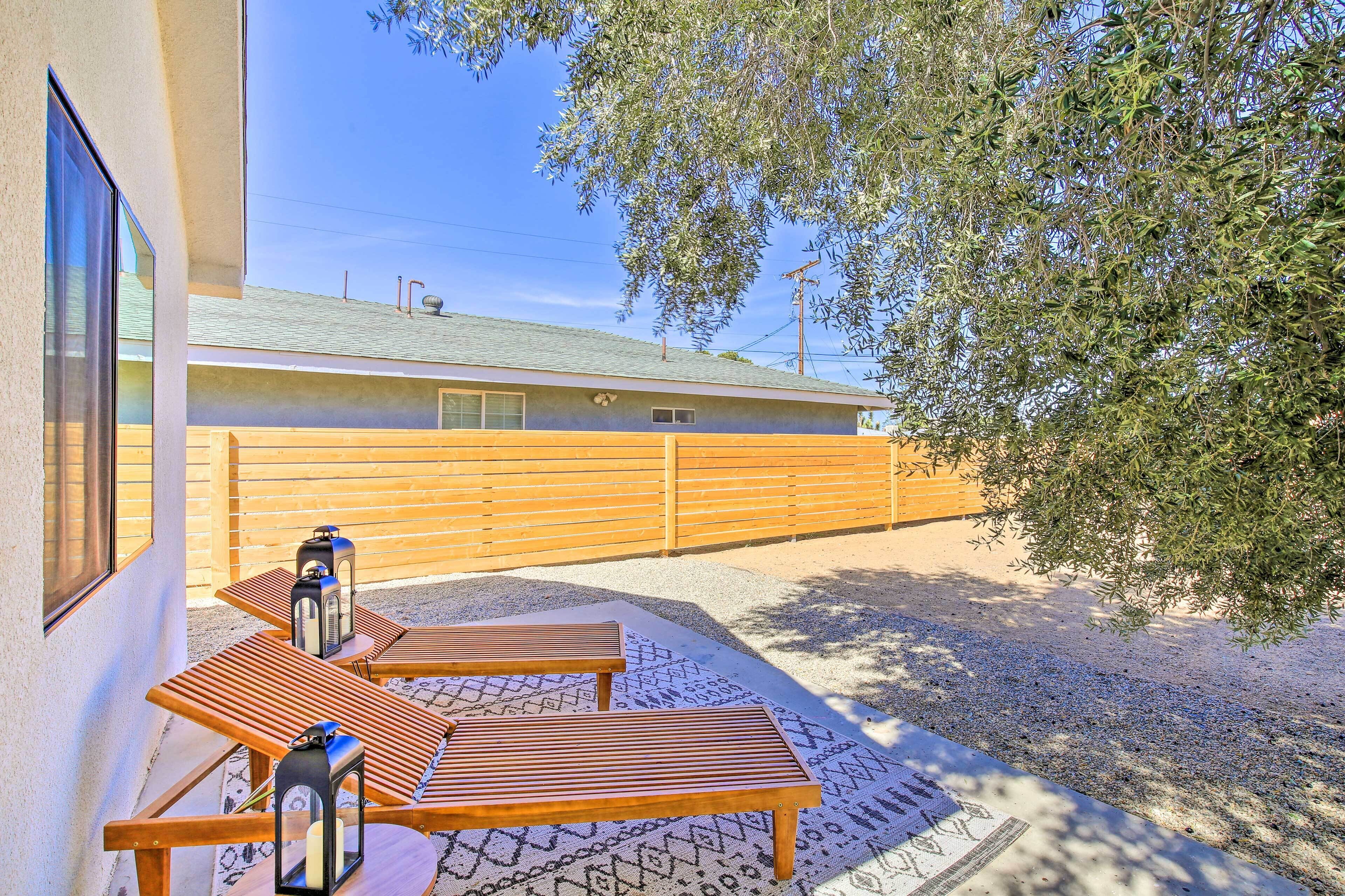 A patio with wooden tables and benches, a lantern, and a view of a building with a wooden fence and trees.