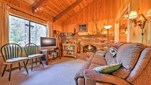 A cozy cabin interior with a fireplace, wooden walls, a sofa, and two chairs. - Big Bear Lake Cabin w/ Deck ~ 7 Mi to Ski Slopes! (Fawnskin)
