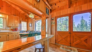 A wooden cabin interior with a kitchen area, dining table, chairs, and large windows offering a view of the forest. - Big Bear Lake Cabin w/ Deck ~ 7 Mi to Ski Slopes! (Fawnskin)
