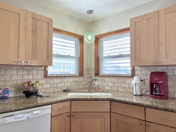 A kitchen with wooden cabinets, a granite countertop, a sink, a coffee maker, and a window with blinds.
