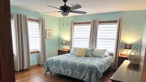 A bedroom with a bed, two bedside tables, a ceiling fan, and large windows with curtains. - IslandSeabreeze-Steps to Beach+1 Night Stays+Great for Cruisers/Families/Couples (Galveston)