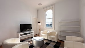A modern living room with a flat-screen TV, white sofas, a round ottoman, and a large window with a view of the city. - Private Unit in Heritage Building in Fitzroy North (Fitzroy North)