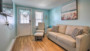 Un salon avec un canapé beige, une petite table et une chaise. On y trouve une porte avec une fenêtre et un tableau accroché au mur.