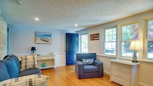 Una sala de estar con un sofá azul, un sillón gris, una mesita de noche blanca con una lámpara y una puerta que da al exterior.
