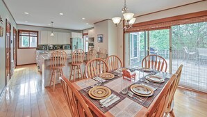 A spacious dining area with a wooden table set for six, surrounded by wooden chairs. A kitchen with modern appliances is visible in the background. - Walk to Beach! Hyannis Family Haven w/ Deck (Hyannis)