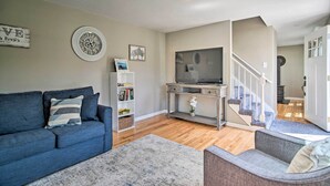 Een woonkamer met een blauwe bank, een grijze fauteuil, een flatscreen televisie, een boekenkast en een trap naar een andere kamer.
