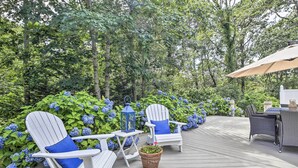 A wooden deck with white Adirondack chairs and a table, surrounded by lush greenery and blue hydrangeas. - ~ 4 Mi to Dtwn: Chatham Home w/ Fire Pit (South Chatham)