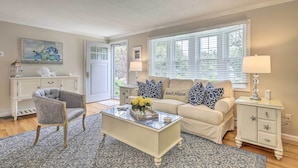 A living room with a beige sofa, a gray armchair, a glass coffee table with a floral centerpiece, and a white sideboard with starfish decorations. - ~ 4 Mi to Dtwn: Chatham Home w/ Fire Pit (South Chatham)
