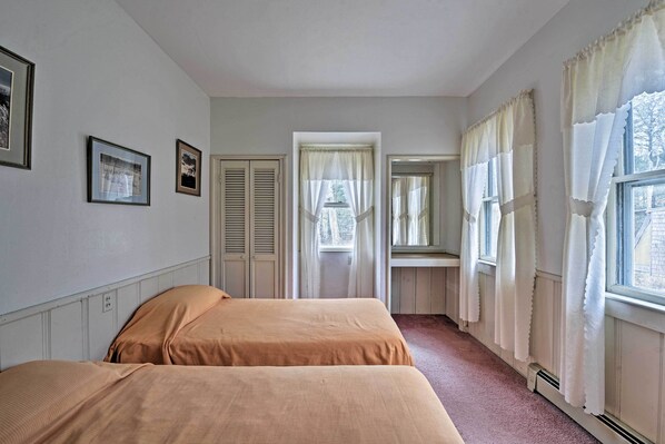 A room with two beds, a window with curtains, a radiator, and framed pictures on the wall.