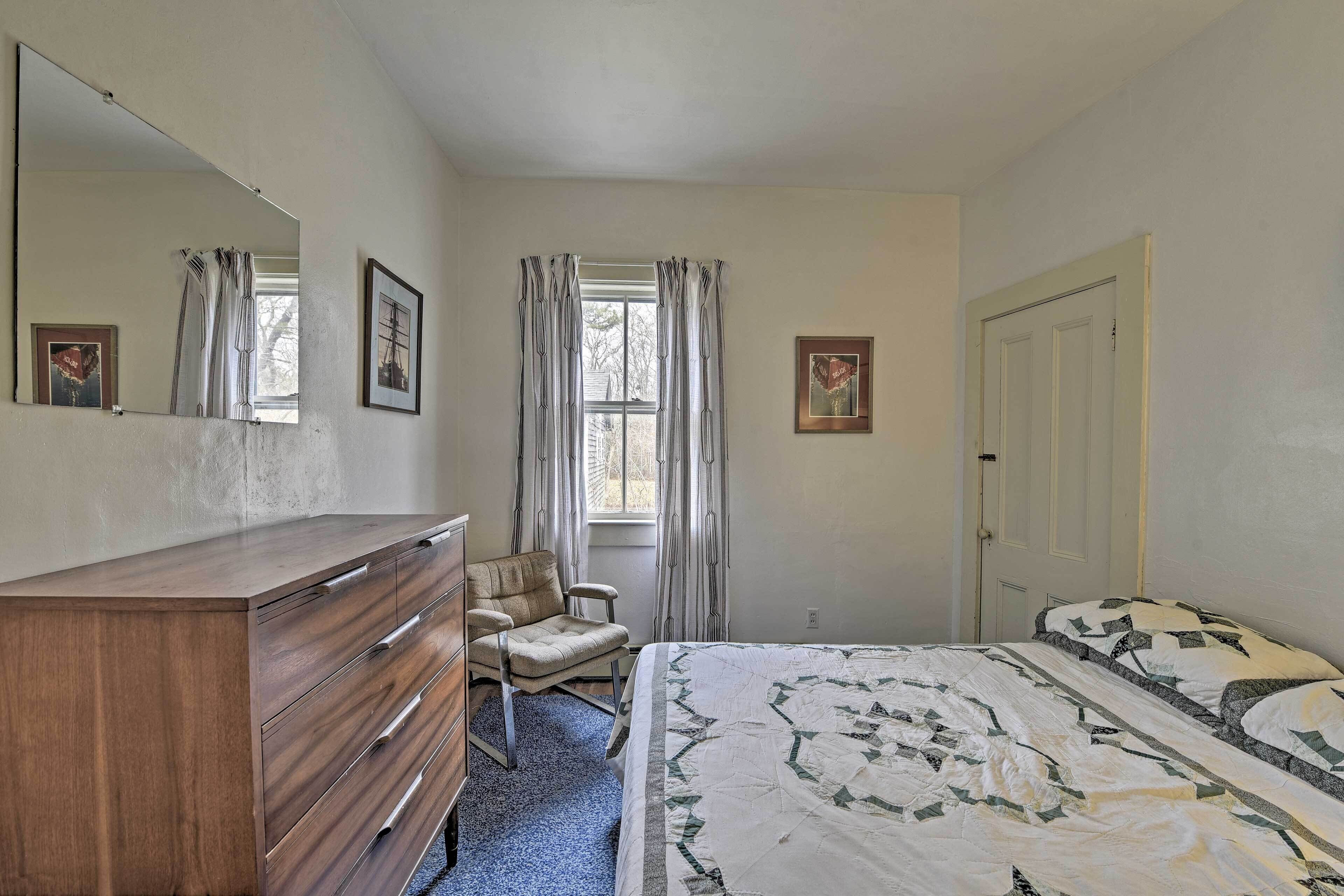 A bedroom with a wooden dresser, a chair, a bed with a patterned quilt, a window with curtains, and a door.