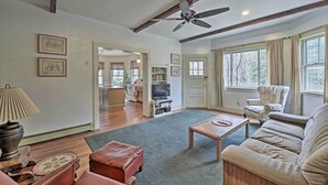 A spacious living room with a ceiling fan, a sofa, a coffee table, and a television. There are framed pictures on the walls and a window with curtains. - Easy Access to Beaches: Vineyard Haven House (Vineyard Haven)