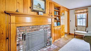 A room with a fireplace, wooden paneling, a brick wall, a wooden cabinet, a chair, and a window. - 2 Mi to Shore: Home w/ BBQ & Beach Gear in Mashpee (Mashpee)