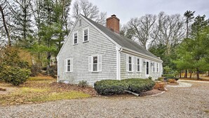 A single-story house with a gabled roof and a brick chimney, surrounded by a gravel driveway and landscaped with shrubs and trees. - 2 Mi to Shore: Home w/ BBQ & Beach Gear in Mashpee (Mashpee)