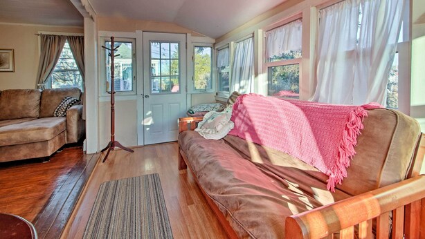 A cozy living room with a brown sofa, a wooden chair, and a wooden floor. There is a door with a coat rack and a window with curtains. - Steps to Cape Cod Canal: Pet-friendly Home (Buzzards Bay)