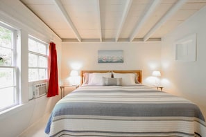 A bedroom with a large bed, two bedside lamps, a painting on the wall, and a window with curtains. - Cozy Stockbridge Cabin - Walk to Beach & Lake! (Stockbridge)