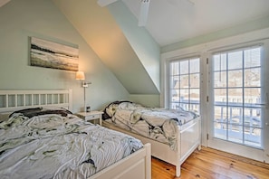 A bedroom with two beds, a nightstand, a lamp, and a window with blinds. - Ocean View + Beach Access: Cape Cod Townhome (Mashpee)