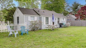 A single-story house with a white exterior, a front porch with two Adirondack chairs, a picnic table, and a barbecue grill. - 1 Mi to Ferry: 'sea Street Cottage' in Hyannis (Hyannis)