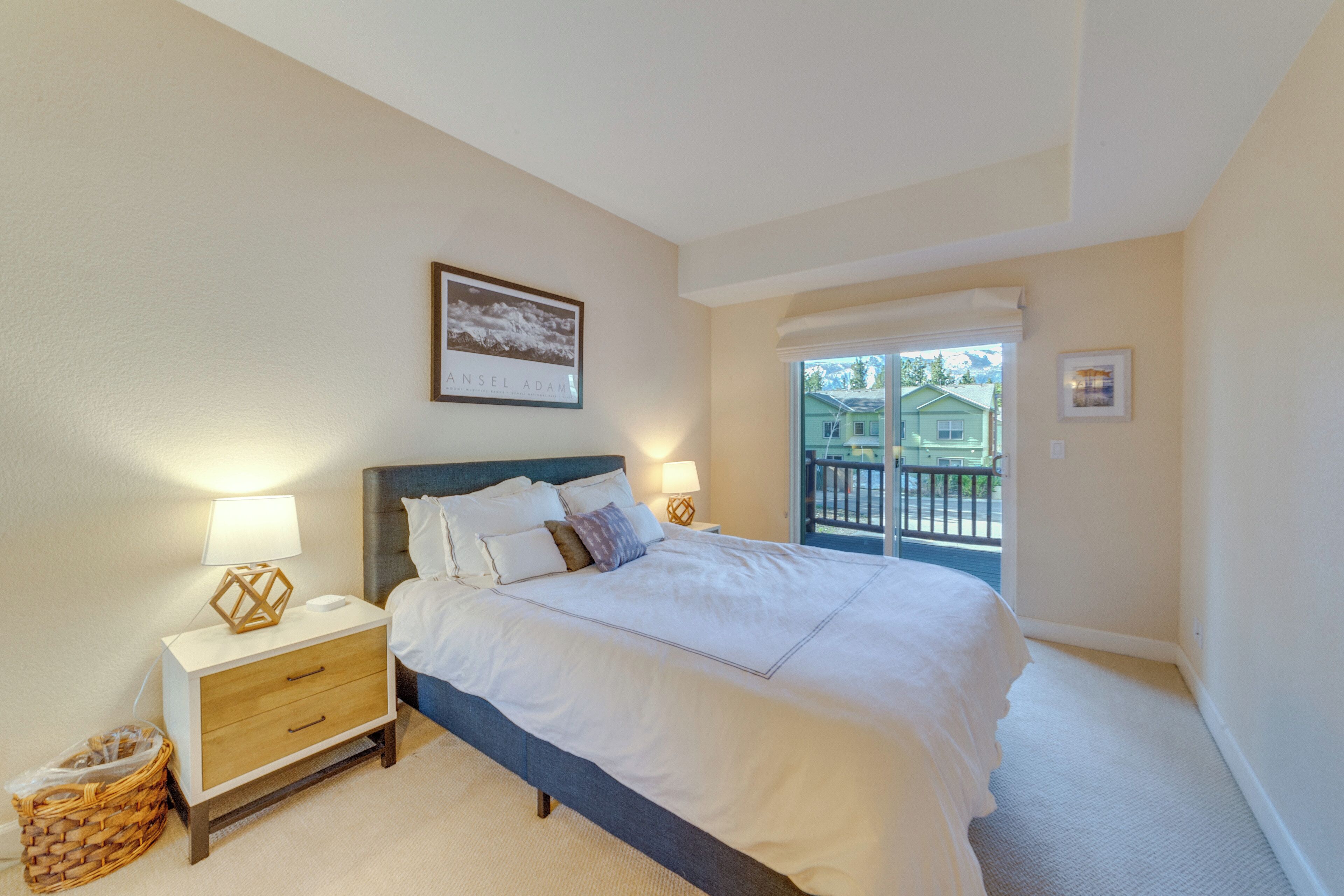 A bedroom with a bed, nightstands, a framed picture, and a view of the outside.