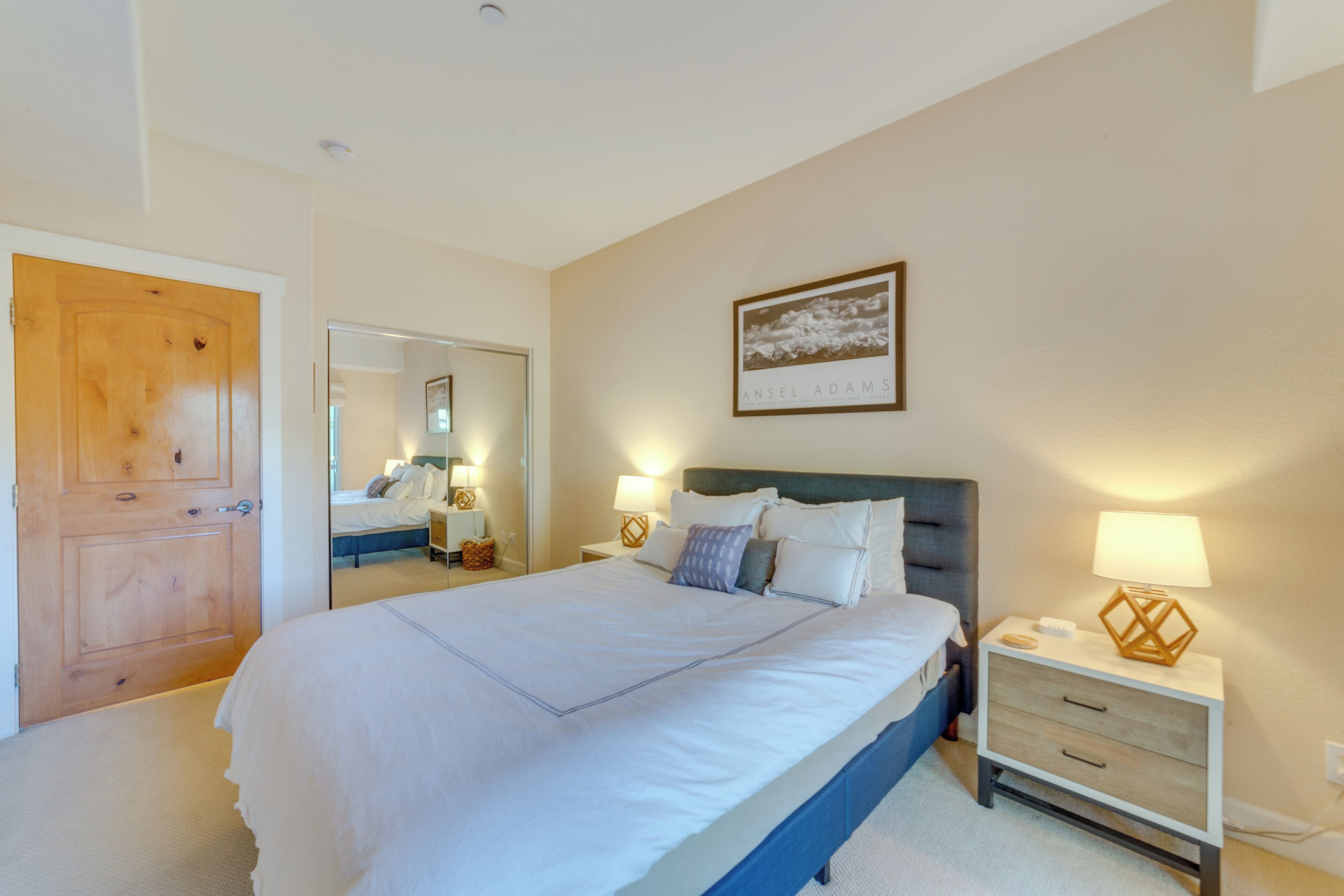 A bedroom with a large bed, a wooden door, a mirror, a bedside table with a lamp, and a framed picture on the wall.