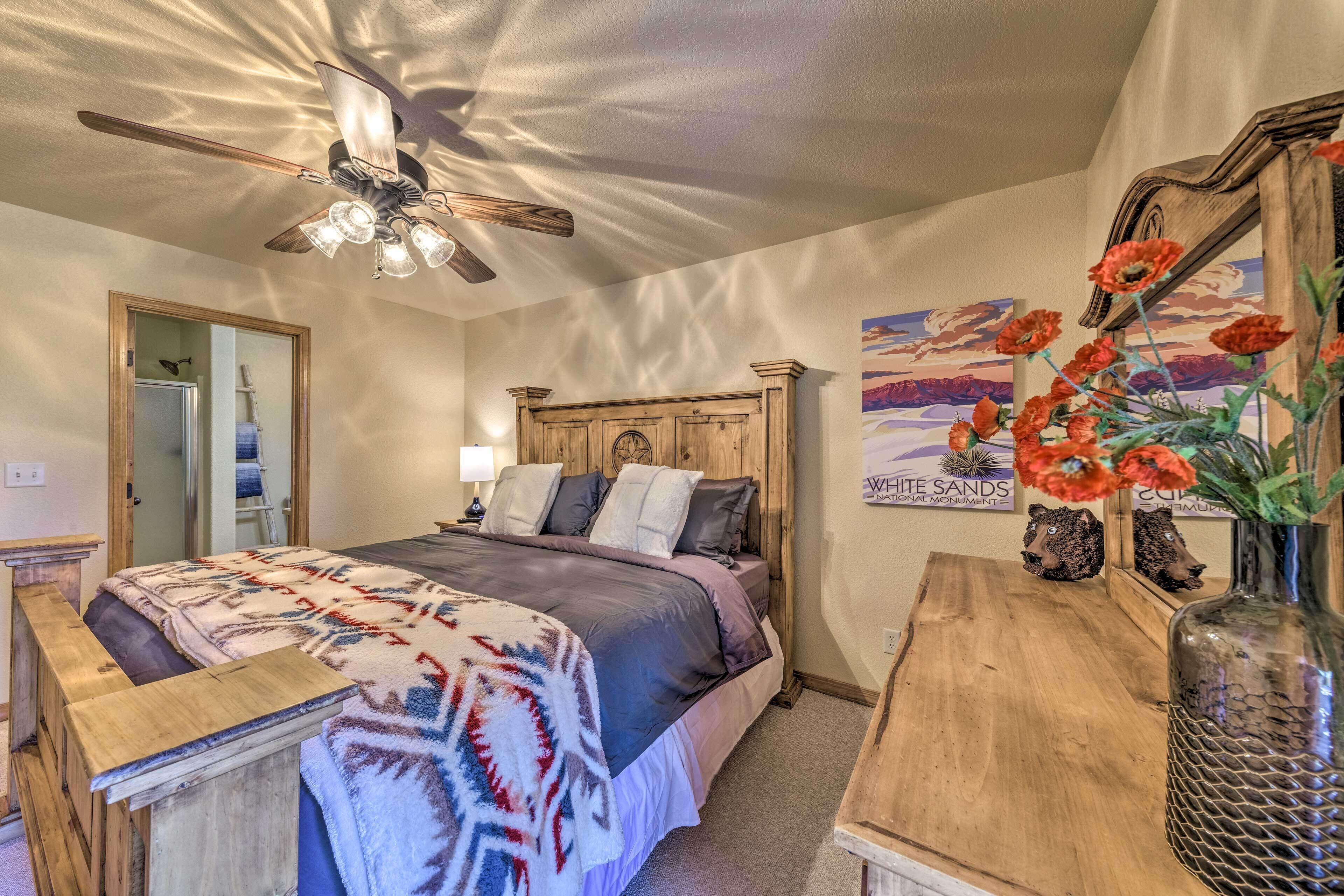 A bedroom with a bed, a wooden headboard, a ceiling fan, a nightstand, a lamp, a mirror, a painting, and a vase with flowers.