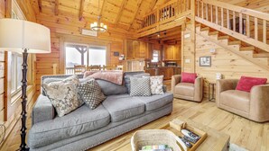 A cozy log cabin interior with a grey sofa, beige armchairs, wooden floors, a staircase, and a kitchen area. - Elevated Cabin Bordering Smoky Mountain Nat'l Park (Bryson City)