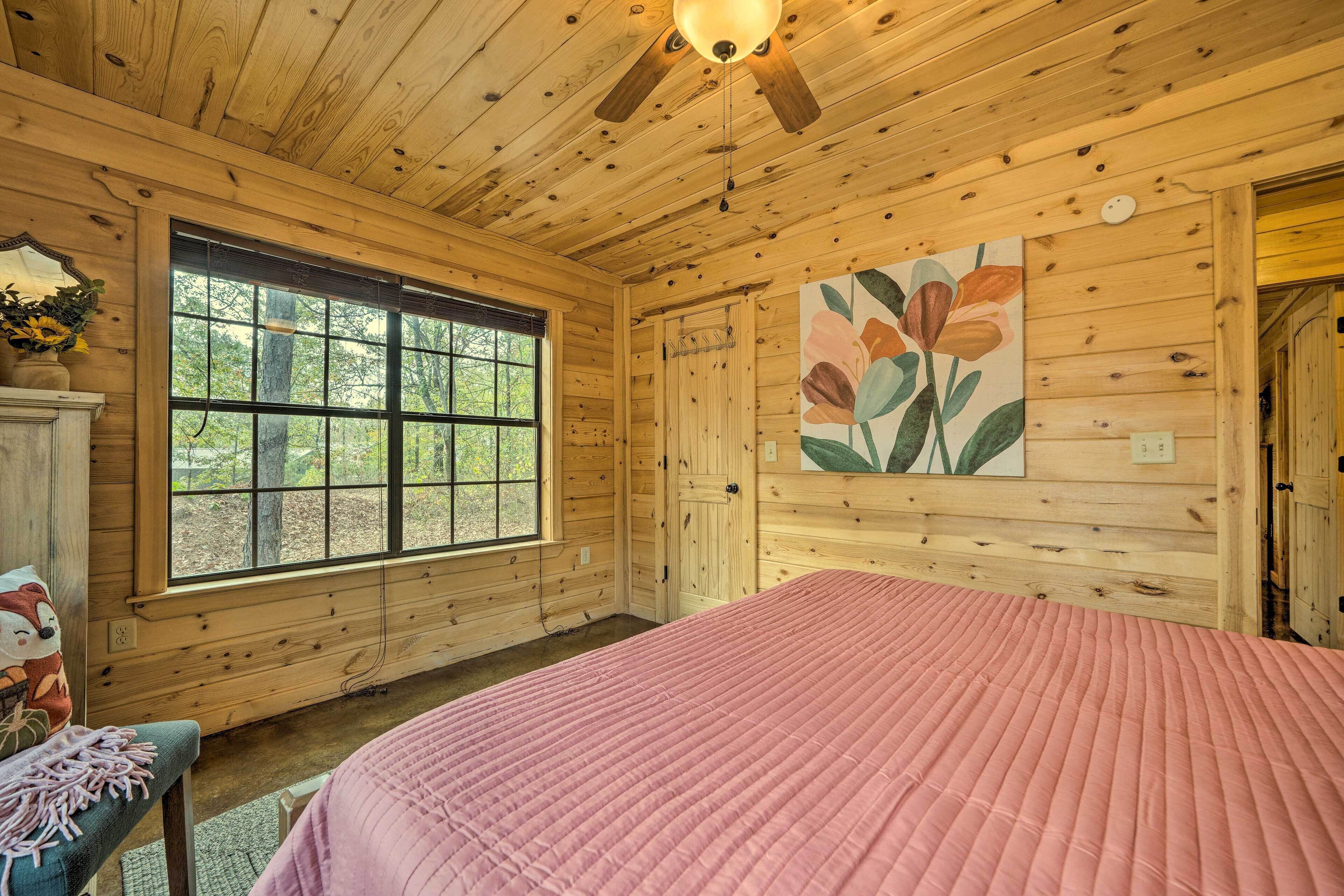 A wooden cabin bedroom with a large window, a bed with a pink quilt, and a ceiling fan.