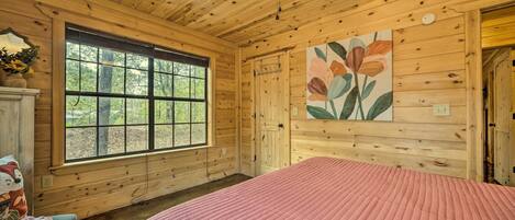 A wooden cabin bedroom with a large window, a bed with a pink quilt, and a ceiling fan.