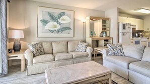 A living room with a beige sofa, patterned pillows, a coffee table, a lamp, and a framed floral artwork.