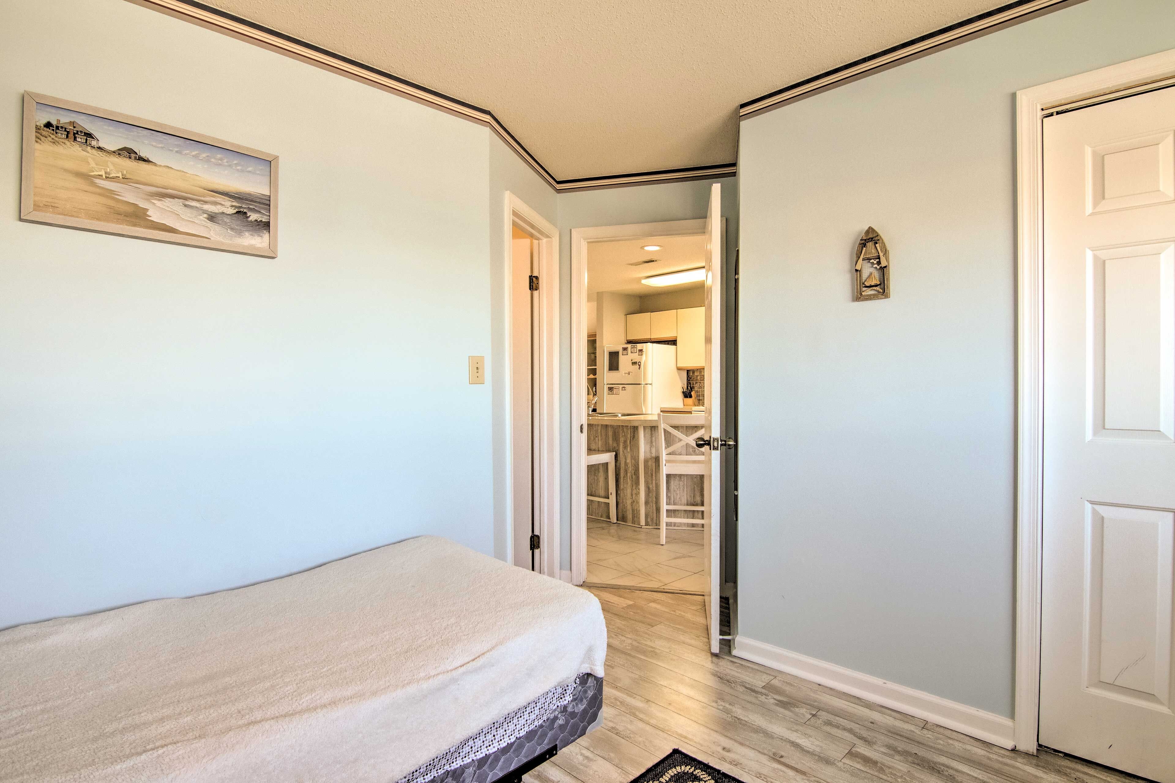 A bedroom with a bed, a painting, and a view into the kitchen.