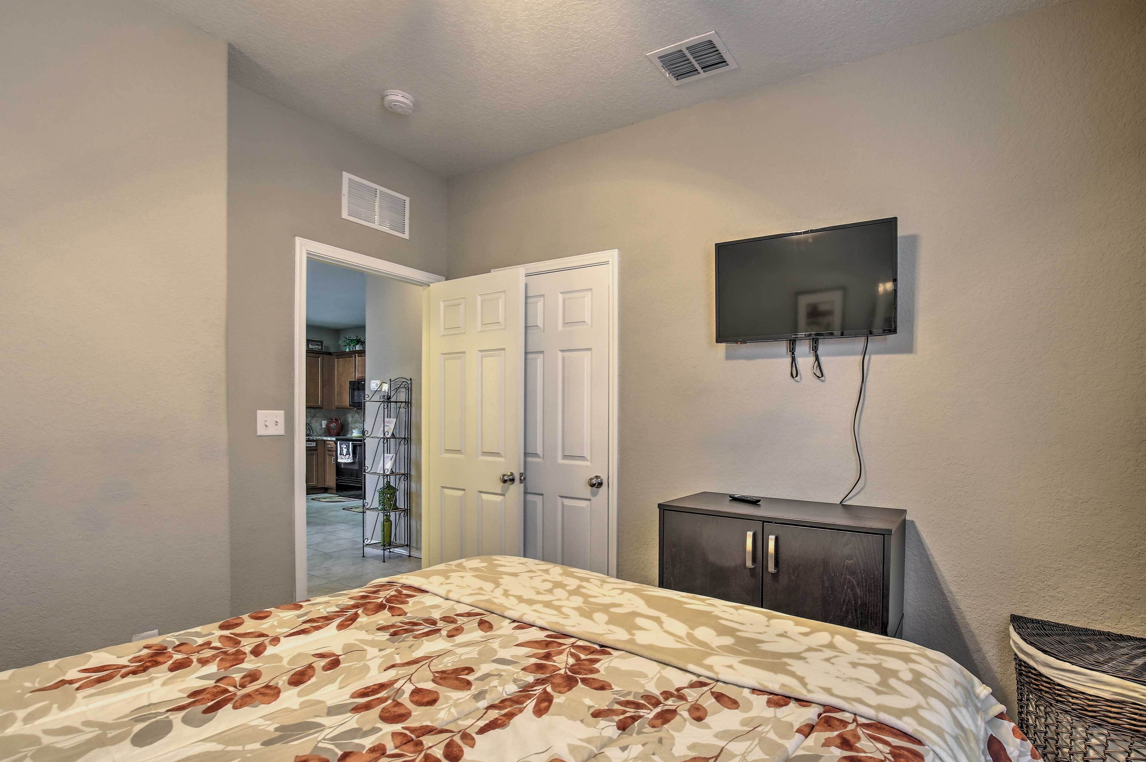A bedroom with a bed, a TV mounted on the wall, a dark cabinet, and a door leading to another room.