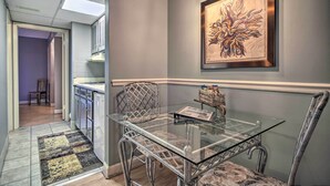 A modern kitchen with a glass dining table and metal chairs, a framed artwork on the wall, and a view into the adjacent dining area.