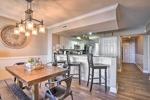 A dining area with a wooden table and chairs, a chandelier, and a kitchen in the background.