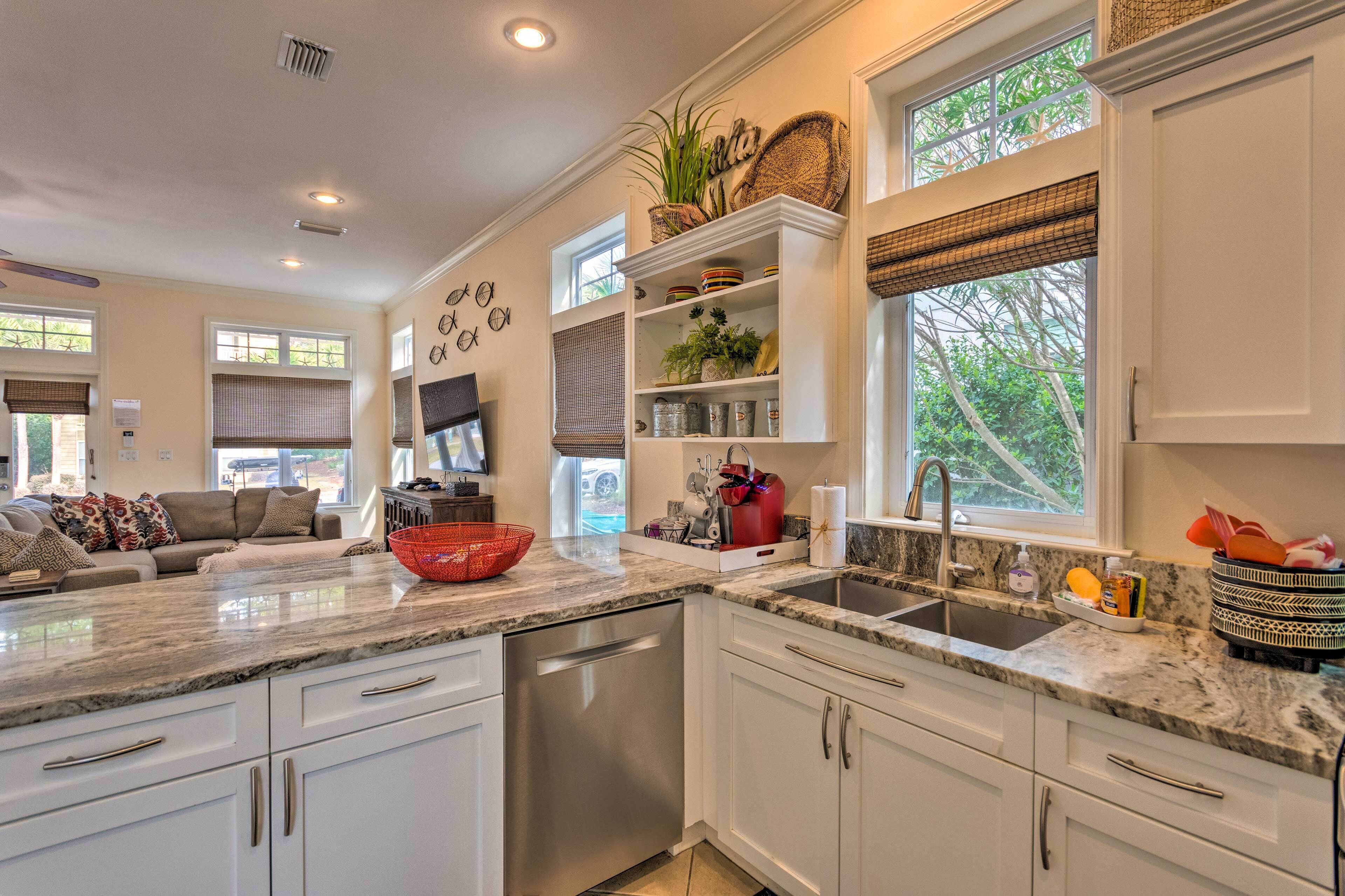 A modern kitchen with white cabinetry, a granite countertop, and a built-in dishwasher. There is a window with a view of greenery outside, and a dining area with a couch and a television.