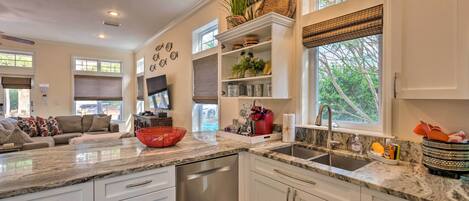 A modern kitchen with white cabinetry, a granite countertop, and a built-in dishwasher. There is a window with a view of greenery outside, and a dining area with a couch and a television.