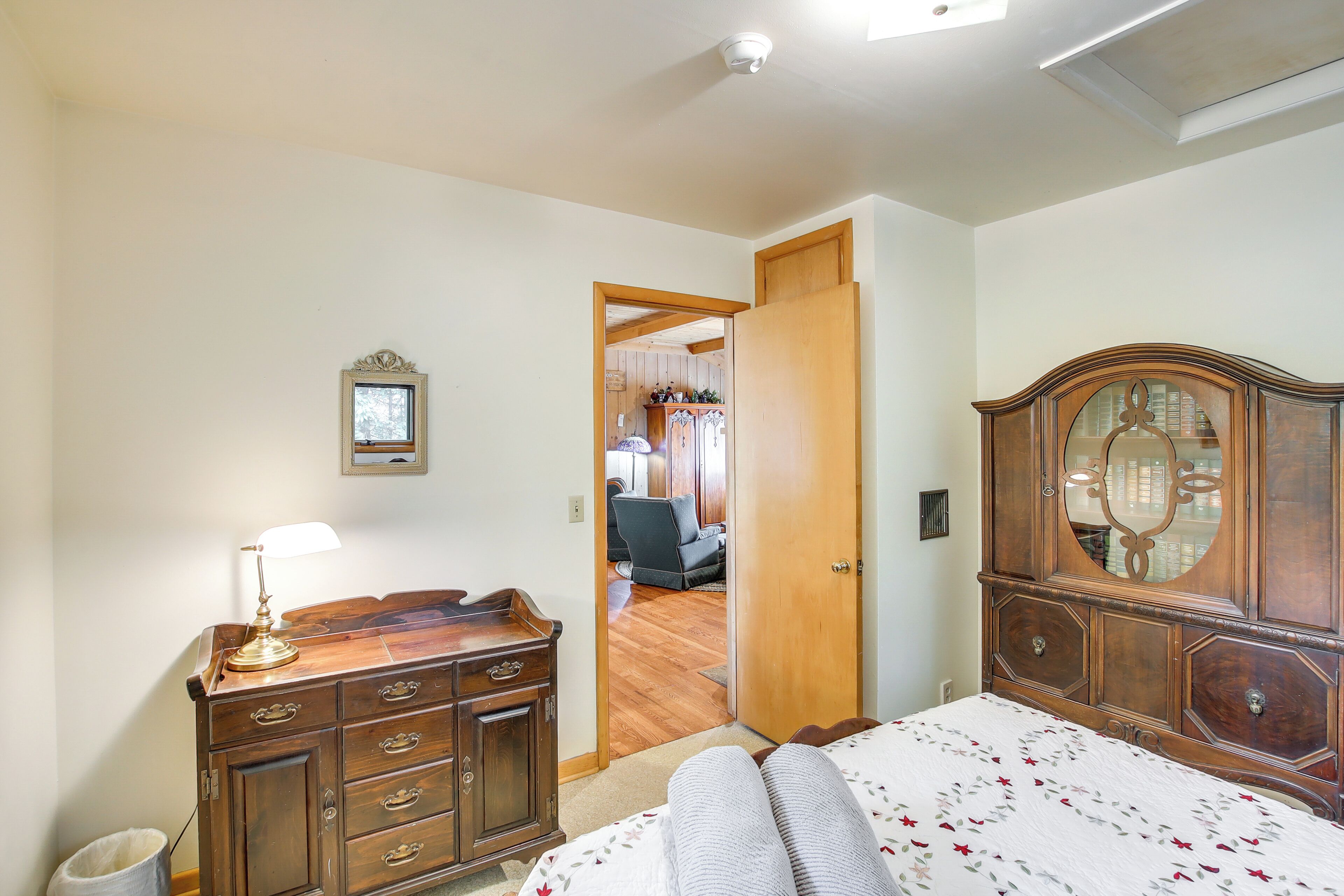 A bedroom with a wooden dresser, a bed with a floral bedspread, a mirror above the dresser, and a door leading to another room.