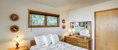Ein Schlafzimmer mit Bett, Nachttisch, Lampe, Kommode, Spiegel und einem Fenster mit Blick auf Bäume.