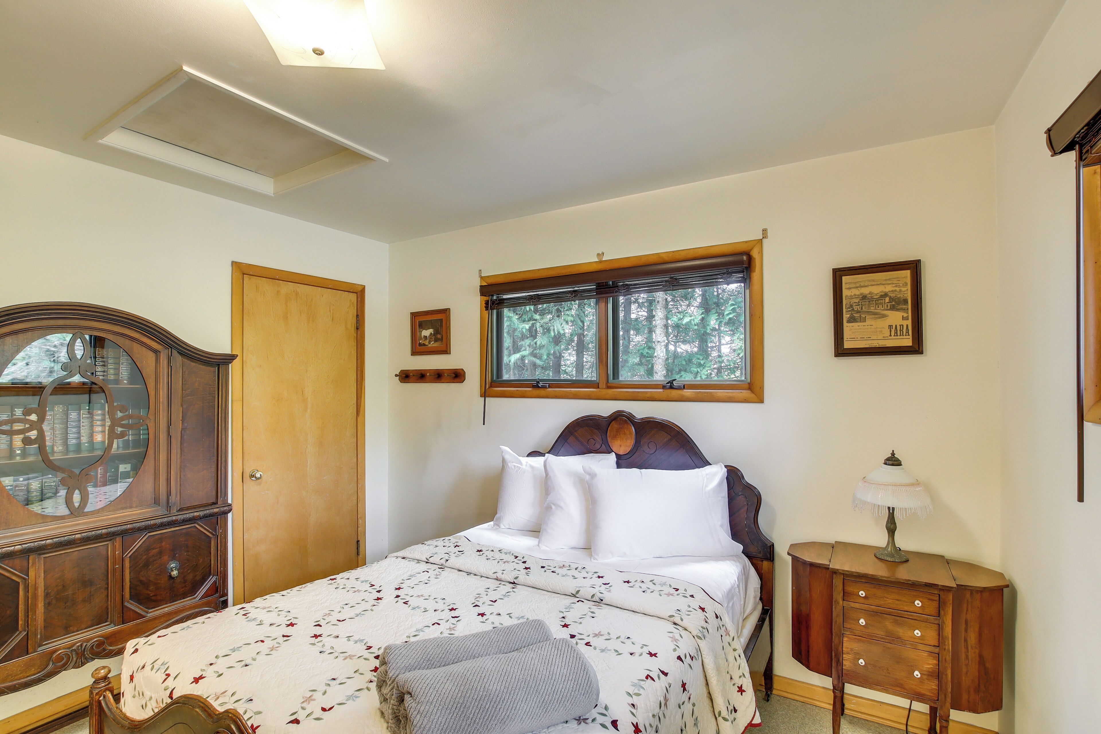A bedroom with a bed, a wooden dresser, a nightstand, a window with a view of trees, and a ceiling light.
