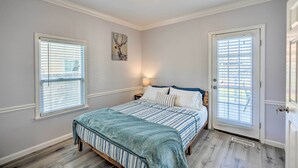 A bedroom with a bed, two windows with blinds, a nightstand, a wall-mounted light, and a door.