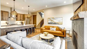 A modern living room with a sofa, coffee table, and a fireplace. There is a kitchen area with wooden cabinets and a stainless steel refrigerator. A staircase is visible in the background. - Winter Park Vacation Rental ~ 4 Mi to Slopes (Winter Park)